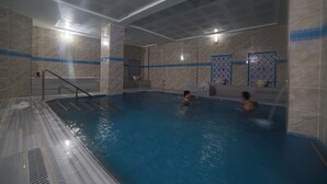 An indoor swimming pool with two people swimming, tiled walls, and a shower area. - ŞAHİN TERMAL OTEL VE SPA (Kutahya)