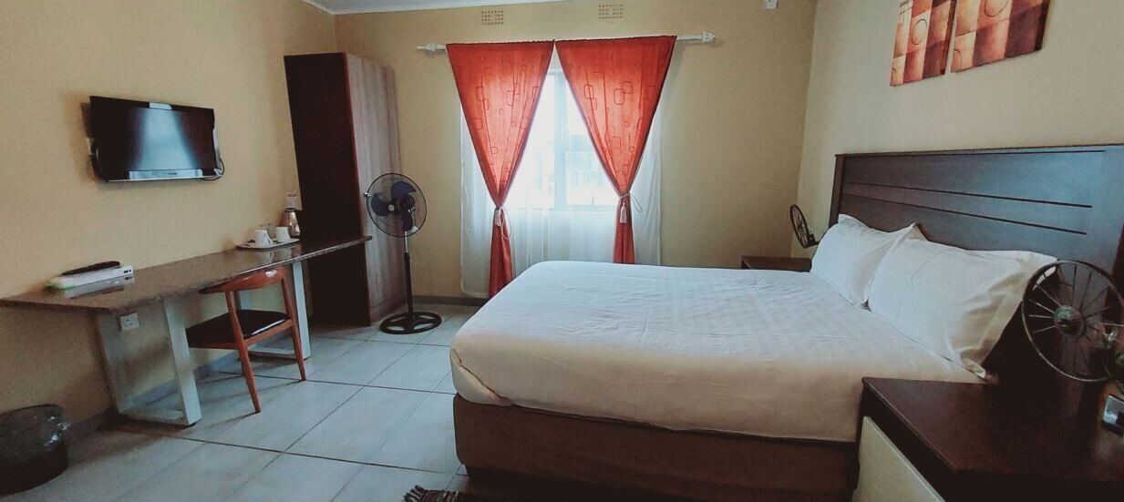 A hotel room with a bed, a desk, a TV, a fan, and a window with curtains.