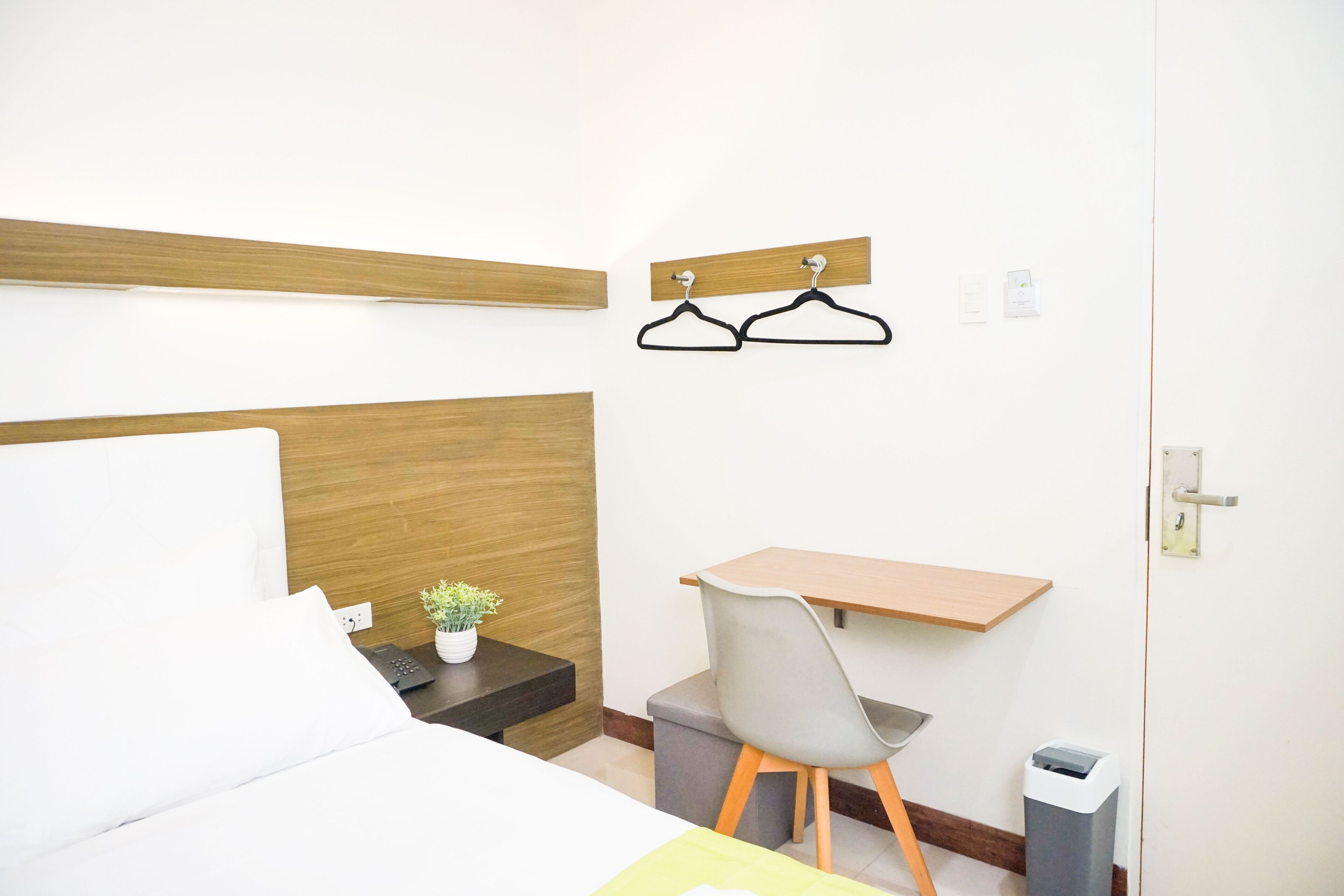 A hotel room with a bed, a desk, a chair, a small plant, and a trash bin.