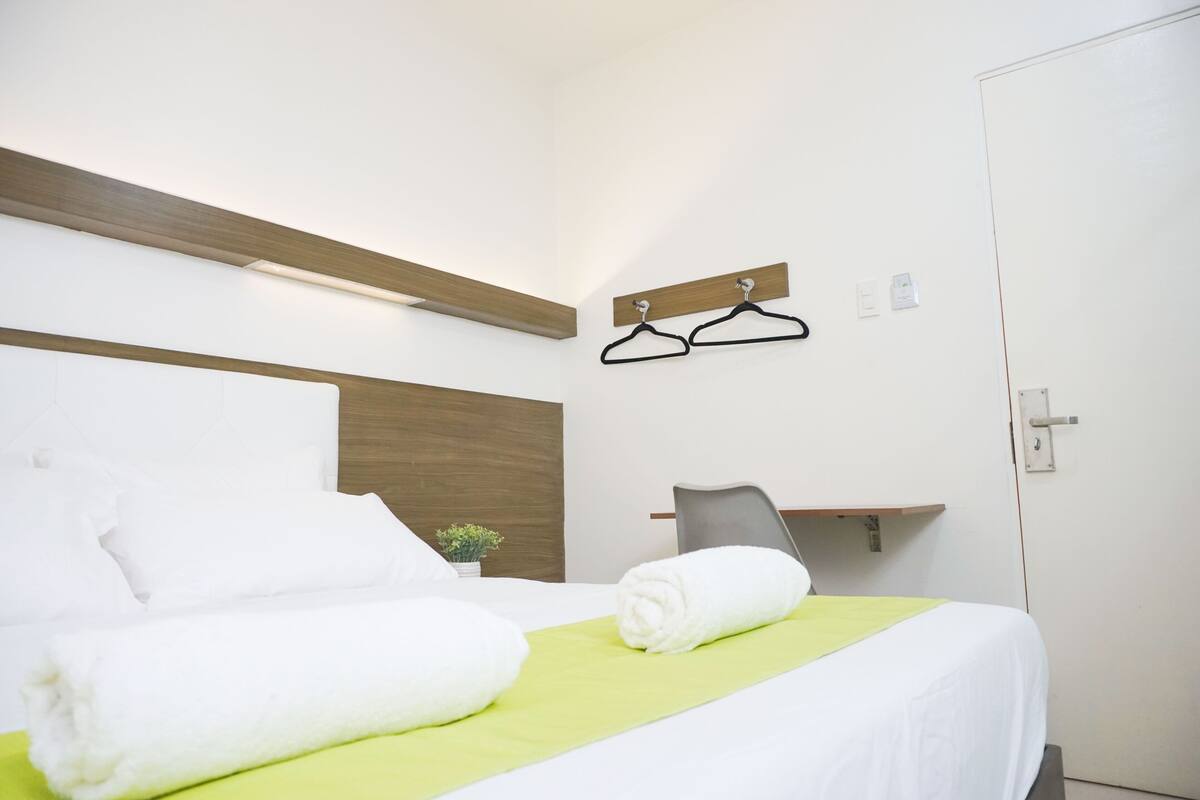 A hotel room with a neatly made bed, a green bedspread, a wooden headboard, a small plant, and a wall-mounted coat rack.