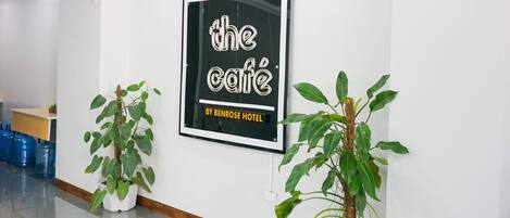 Un vestíbulo de hotel moderno y limpio con un gran cartel enmarcado que dice "the cafe by benrose hotel" y dos macetas con plantas en el suelo.