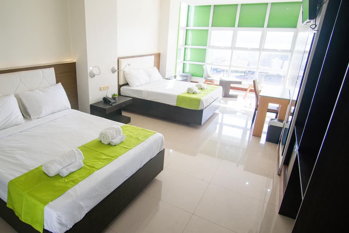A hotel room with two beds, a green accent on the beds, a nightstand with a phone, and a view of the city through large windows.