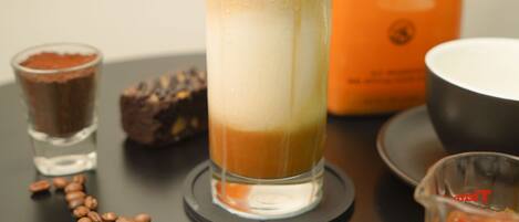 Un verre contenant une boisson à plusieurs couches, une bouteille de sirop au caramel Torani, un petit verre à shooter, un verre doseur et des grains de café éparpillés sur une surface sombre.