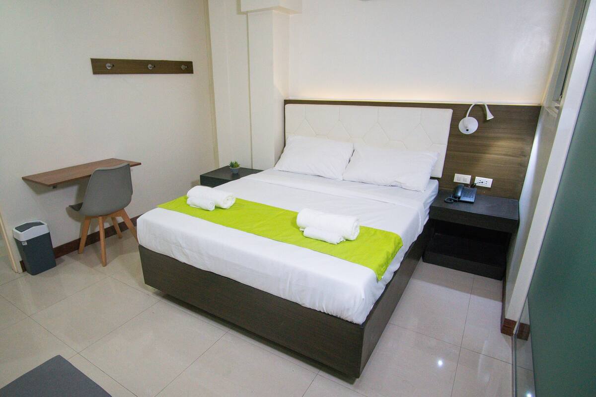 A neatly made bed with a green bedspread, a bedside table with a lamp, a chair, and a small plant.