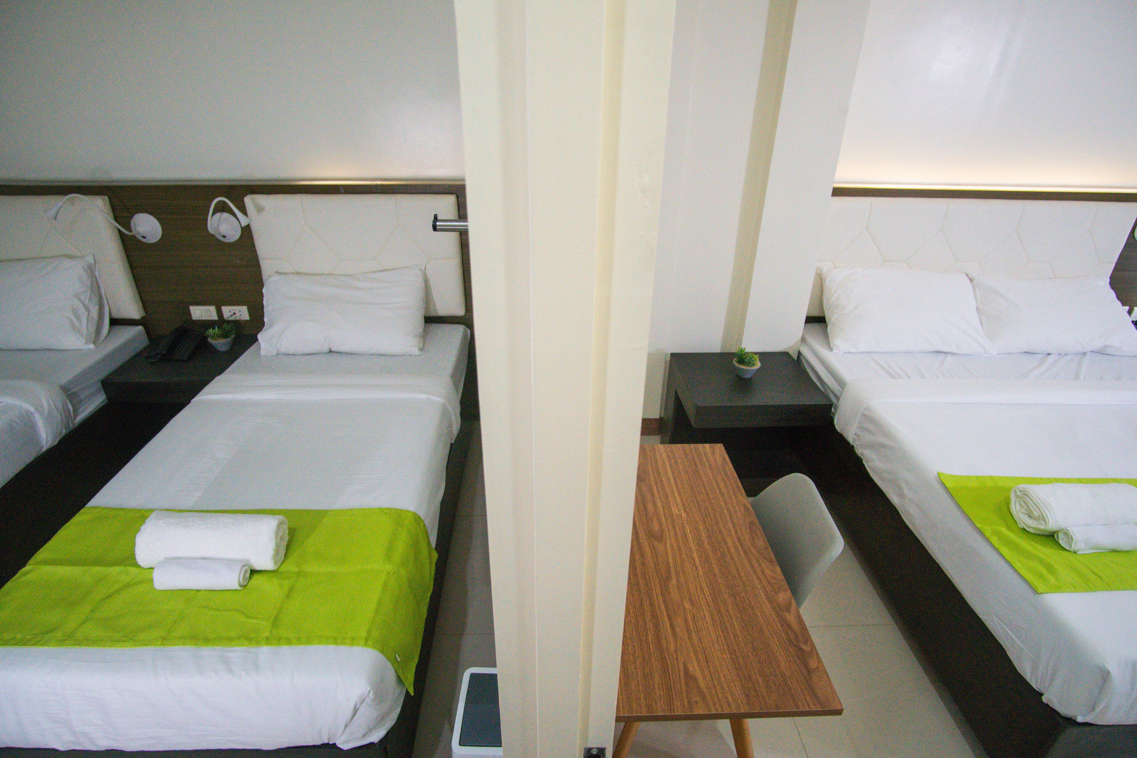 Une chambre d’hôtel avec deux lits, chacun agrémenté de draps blancs et d’une couvre-lit verte. On y trouve une table de chevet en bois, une lampe et une petite plante en pot.