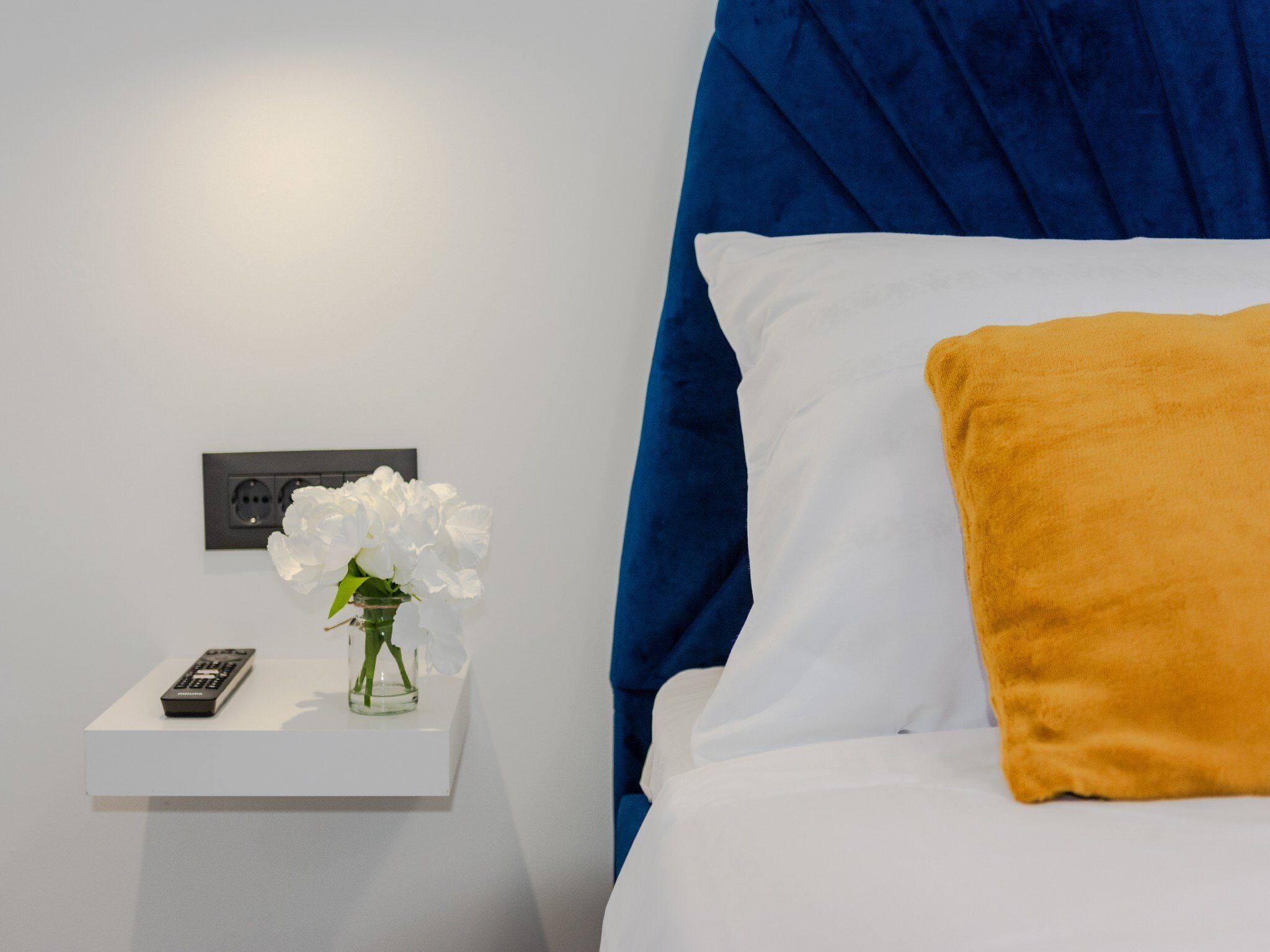 A neatly made bed with white and yellow pillows, a remote control, and a vase with white flowers on a bedside table.