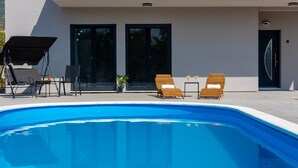 Ett modernt poolområde med en klarblå pool, två träsolstolar, ett litet bord med en krukväxt och en överbyggd sittplats.
