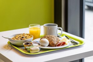 Bandeja de desayuno con cereal, huevos, tostadas y un vaso de zumo de naranja sobre una mesa blanca con bandeja verde.