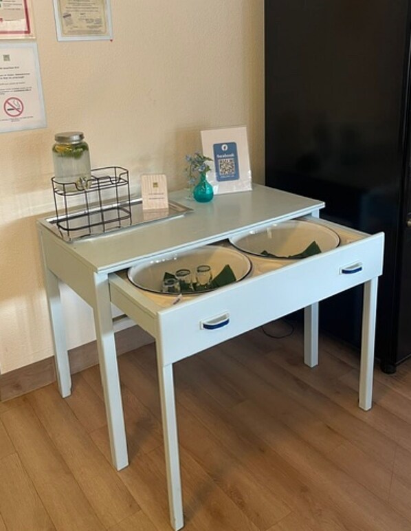 Un petit bureau blanc avec un tiroir, un plateau en métal, un pot, une carte et un code QR sur un mur.
