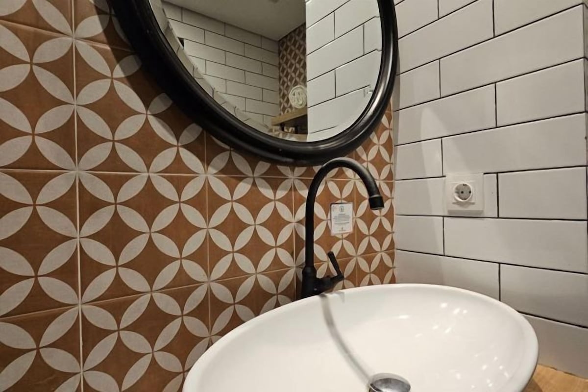 A bathroom with a white sink, a black faucet, and a mirror. The wall has a geometric tile pattern.