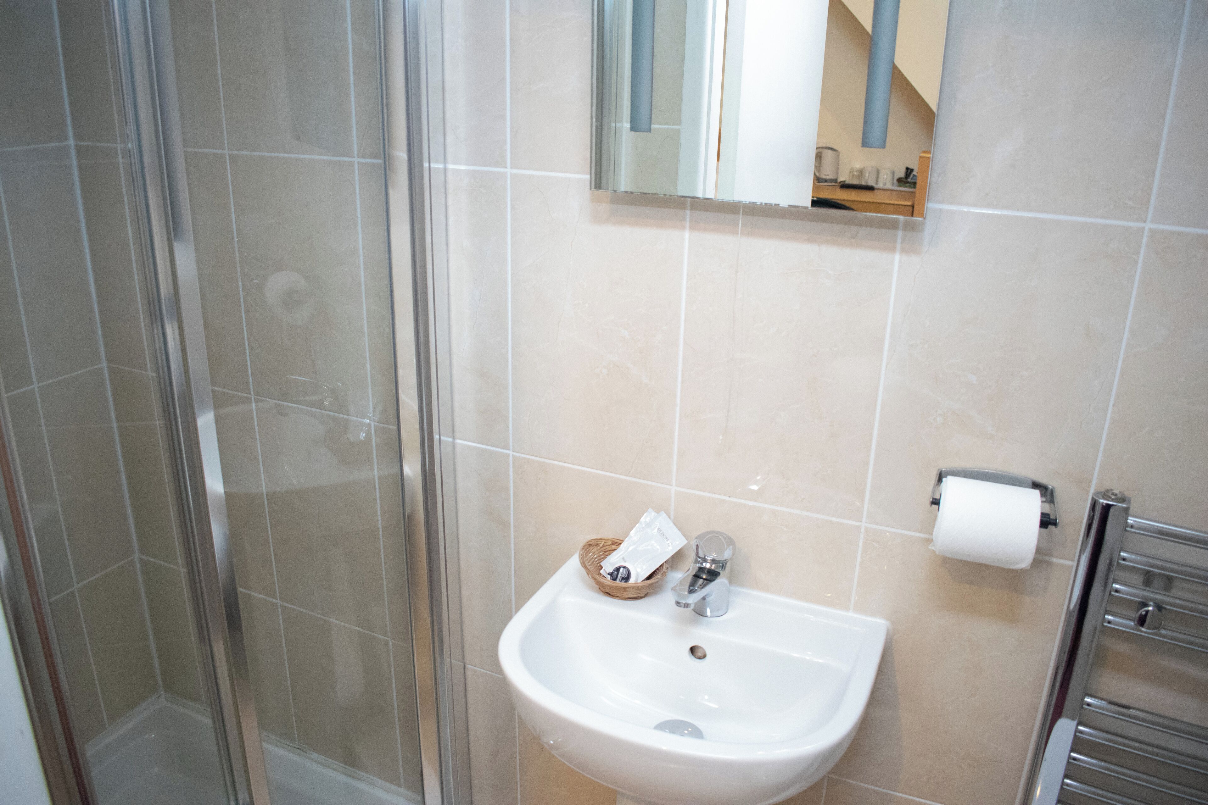 A bathroom with a glass shower enclosure, a sink with a towel dispenser, and a toilet paper holder.