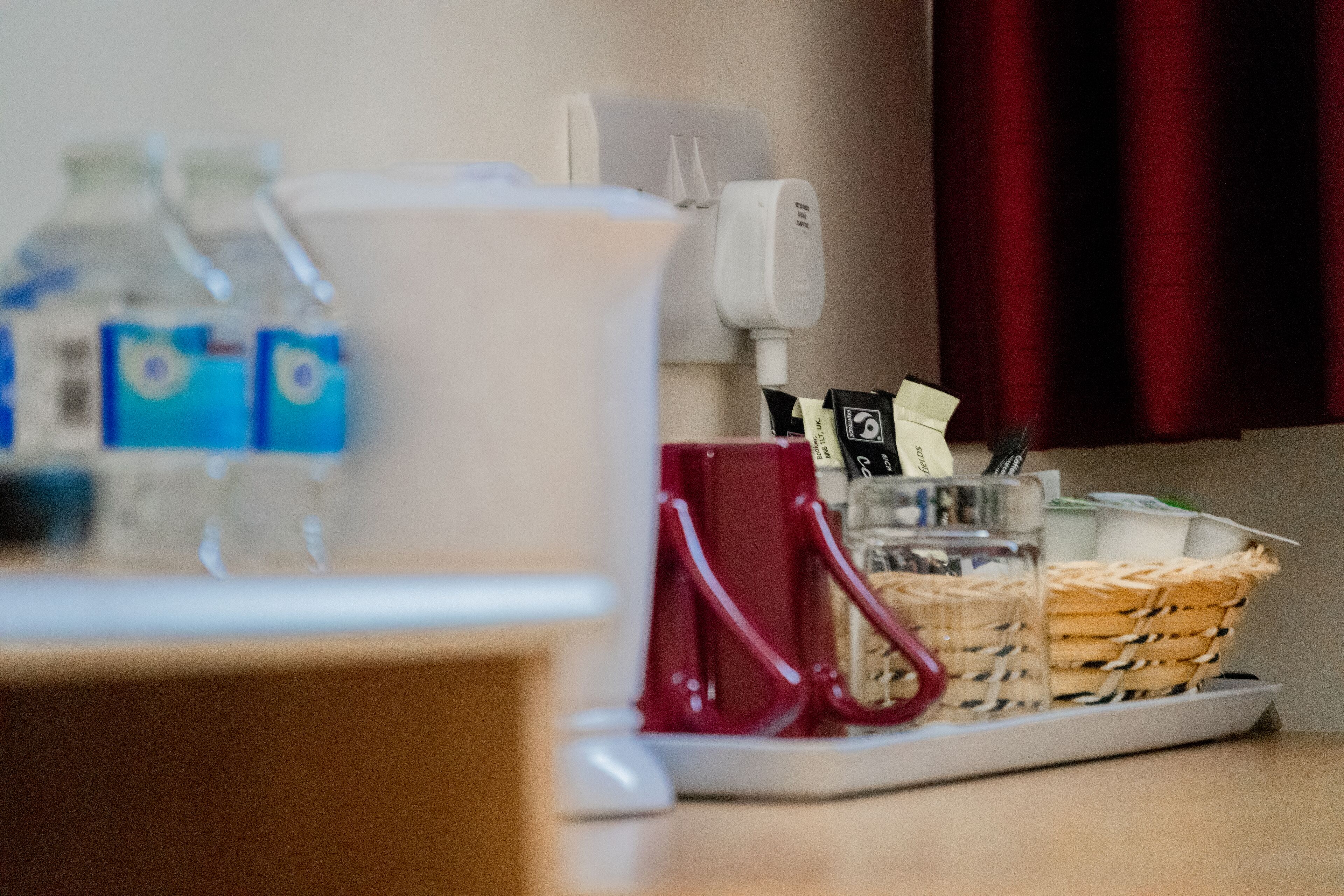 A hotel room with a coffee maker, a red mug, a glass, and a basket of snacks on the table.