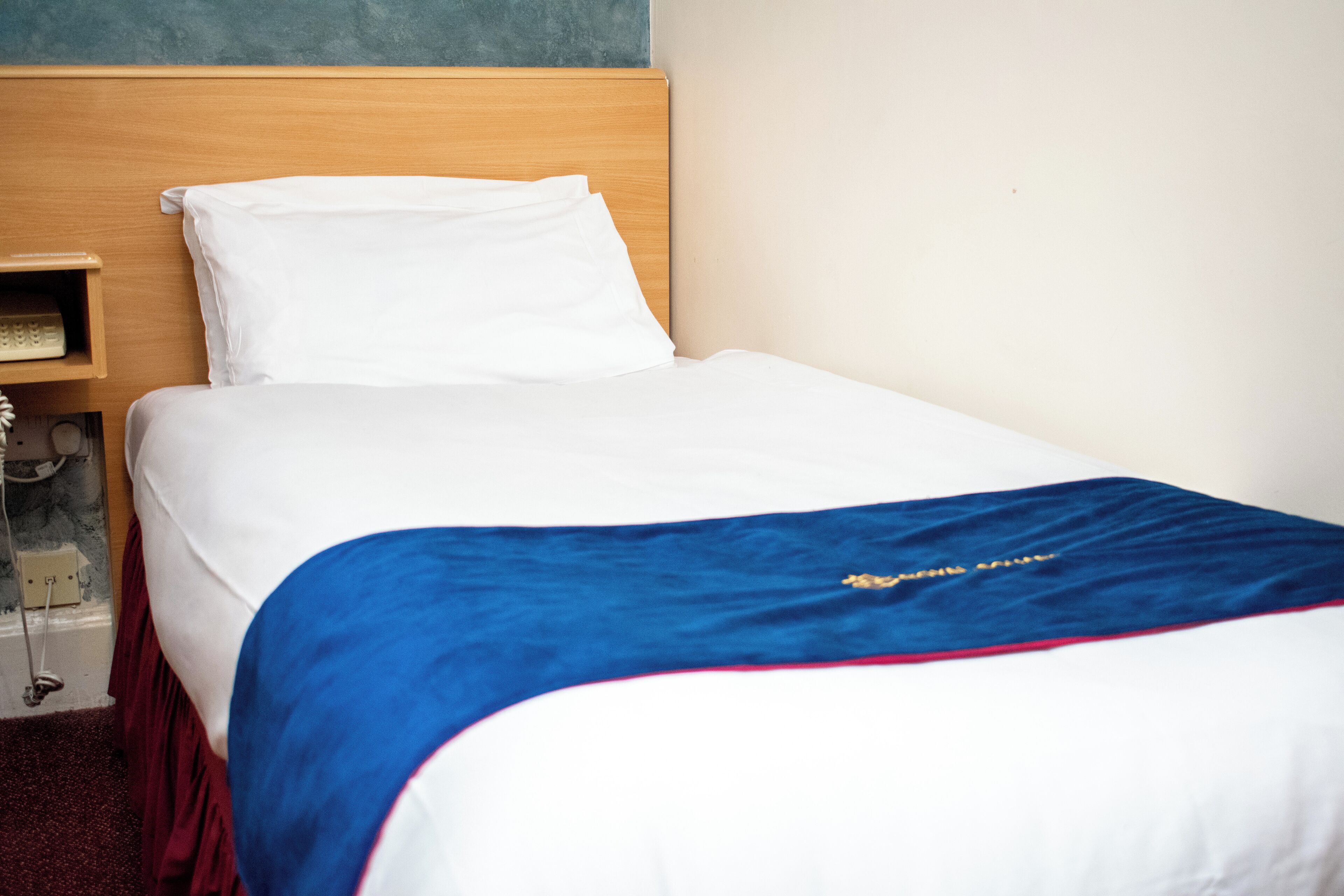 Una cama bien hecha con sábanas blancas y una manta azul, un cabecero de madera y una mesita de noche con una lámpara.