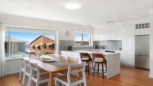 A modern kitchen with white cabinetry, a stainless steel refrigerator, and a dining area with a wooden table and white chairs. - MOON GLOW | Accom Holidays (TERRIGAL)