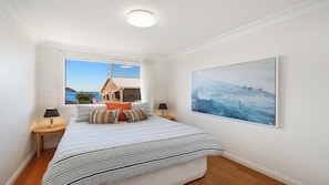 Une chambre à coucher avec un lit, deux tables de chevet, une fenêtre donnant sur un immeuble et la mer, et un grand tableau représentant une vague.