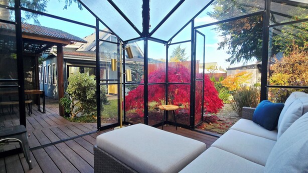 Ein Wintergarten mit Holzterrasse, einer gepolsterten Sitzecke und Blick auf einen Garten mit roten Blättern.