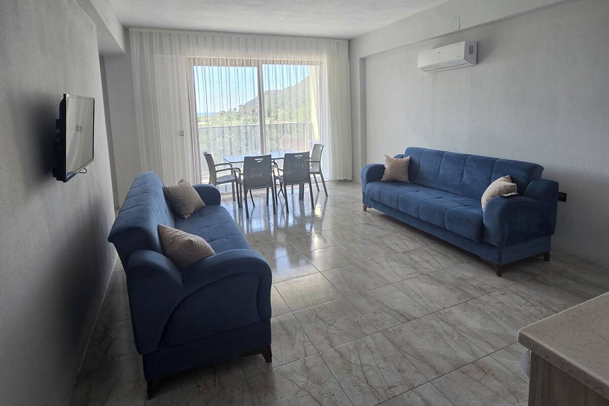 A living room with two blue sofas, a dining table with chairs, a flat-screen TV mounted on the wall, and an air conditioning unit on the wall.