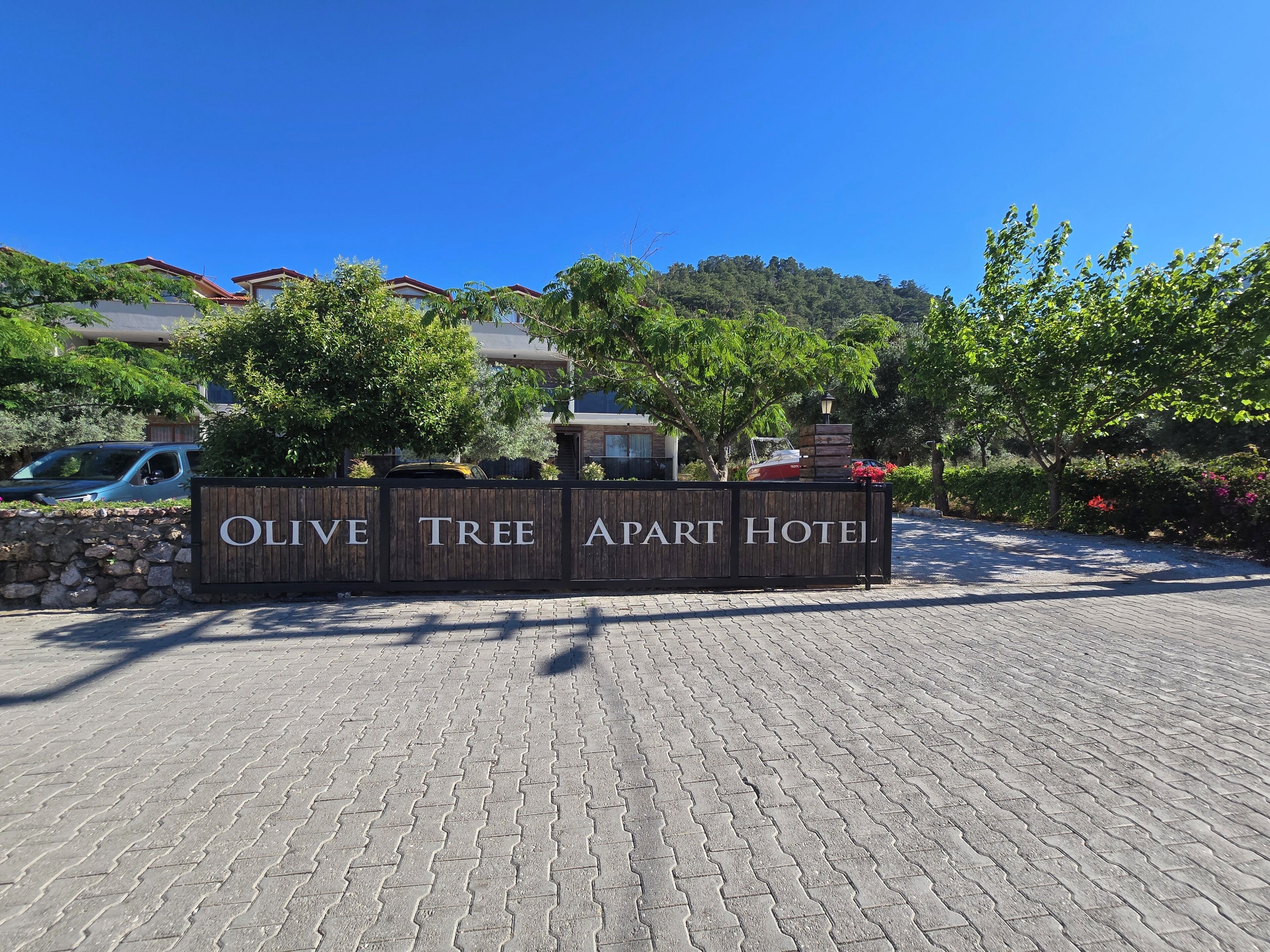 A wooden sign with 'OLIVE TREE APART HOTEL' written on it, set against a backdrop of trees and buildings.