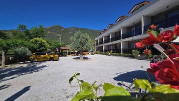 Un hôtel avec une allée gravillonnée, une voiture jaune garée devant et une montagne en arrière-plan.