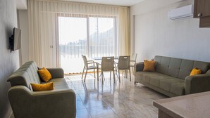 A modern living room with a sofa, chairs, and a dining table. There is a large window with curtains, a flat-screen TV mounted on the wall, and an air conditioning unit. - OLIVE TREE APART HOTEL (Koycegiz)