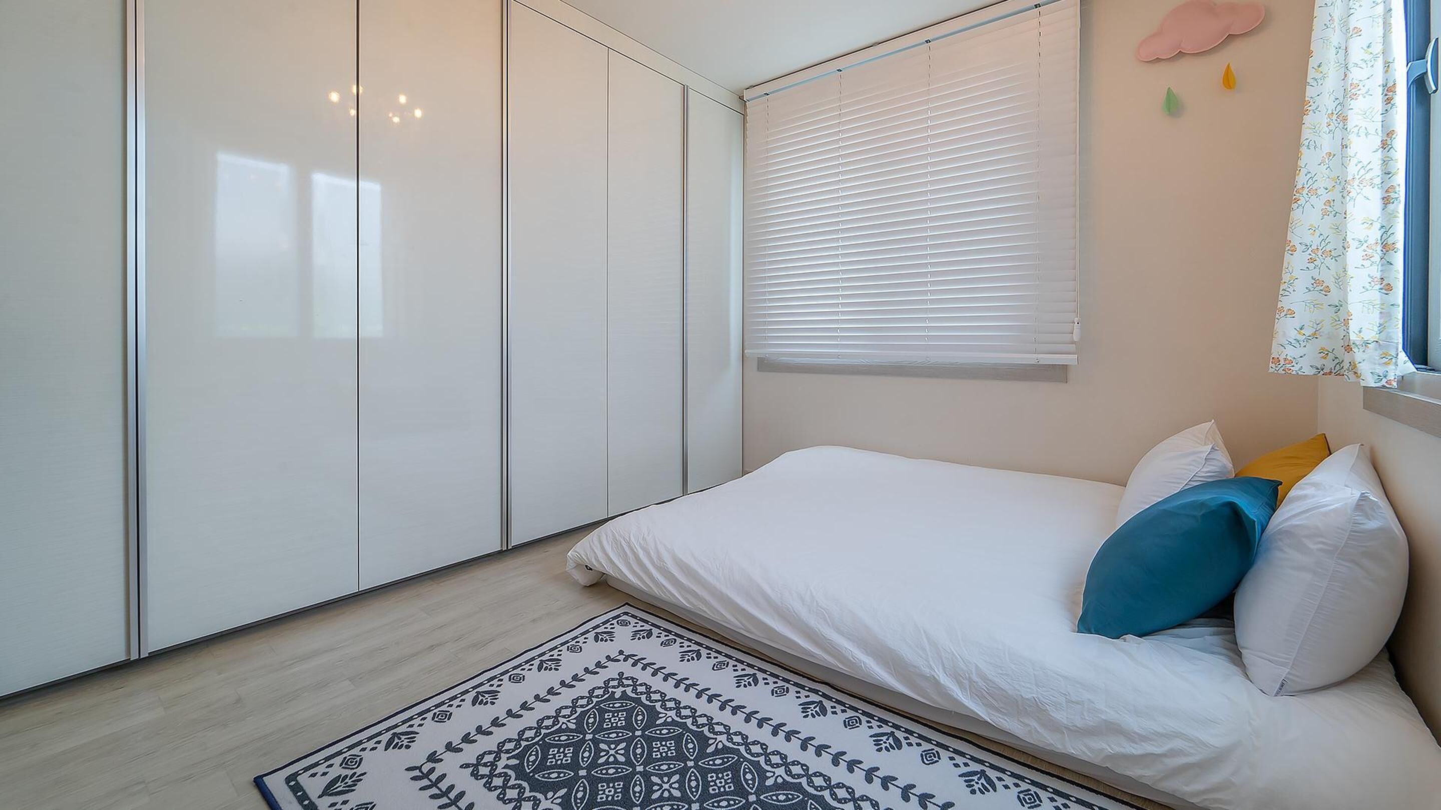 Un dormitorio con cama, almohadas, alfombra estampada, armario y ventana con persianas.