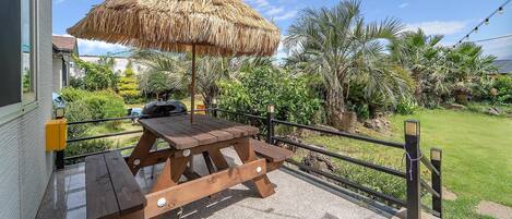 A wooden picnic table with a thatched umbrella, surrounded by a metal fence and lush greenery.