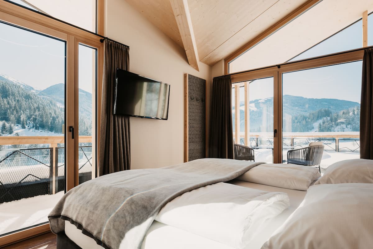 A bedroom with a large bed, a TV mounted on the wall, and a balcony with a view of snow-covered mountains.
