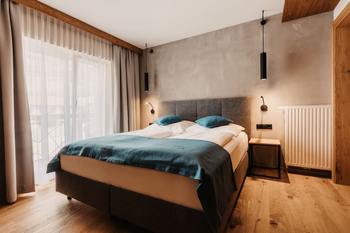 A modern bedroom with a large bed, wooden flooring, and a view of the outdoors through sheer curtains.
