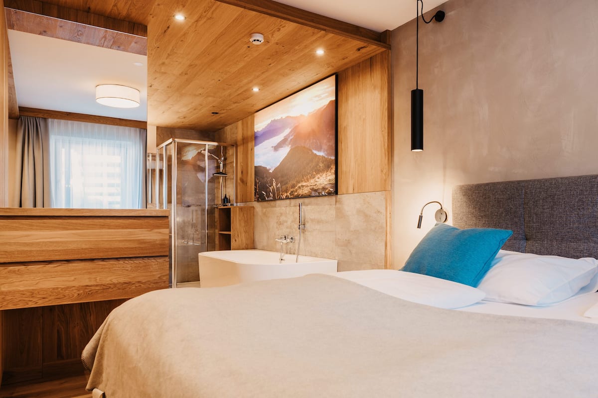 A modern hotel room with a wooden ceiling, a large bed with white and blue pillows, a built-in bathtub, and a large window with a mountain view.