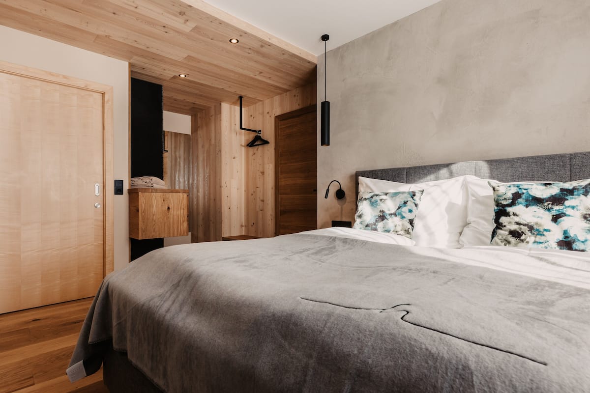 A bedroom with a wooden ceiling, a bed with a grey blanket, a wooden wardrobe, and a concrete wall.