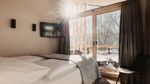 Una habitación de hotel moderna con una cama grande, un televisor de pantalla plana y un ventanal corredero que da a un espacio exterior con terraza de madera y sillas.