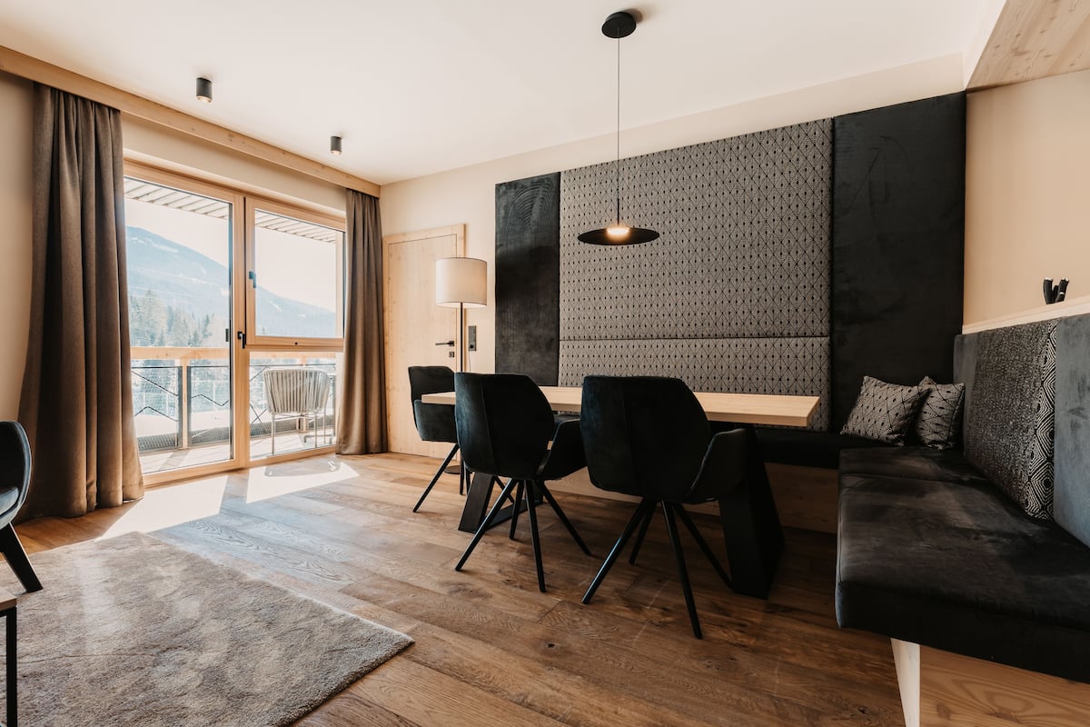 A modern hotel room with a wooden floor, a sofa, a dining table with chairs, and a large window with curtains.