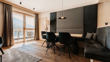 A modern hotel room with a wooden floor, a sofa, a dining table with chairs, and a large window with curtains.