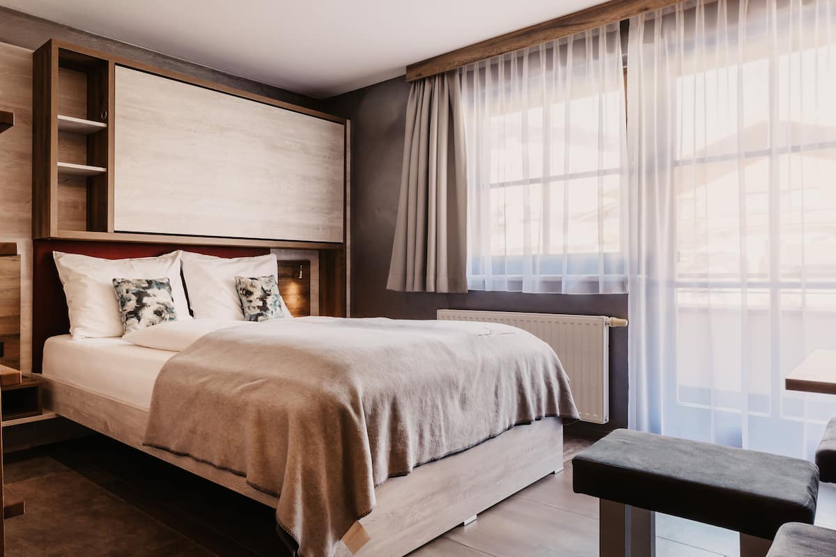 A modern bedroom with a large bed, a wooden headboard, a bedside table, and a window with sheer curtains.