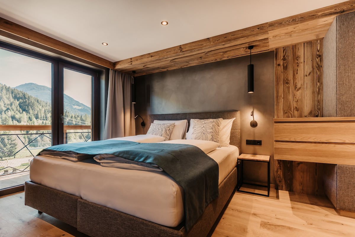 A modern bedroom with a large bed, wooden headboard, and a view of a mountainous landscape through large windows.