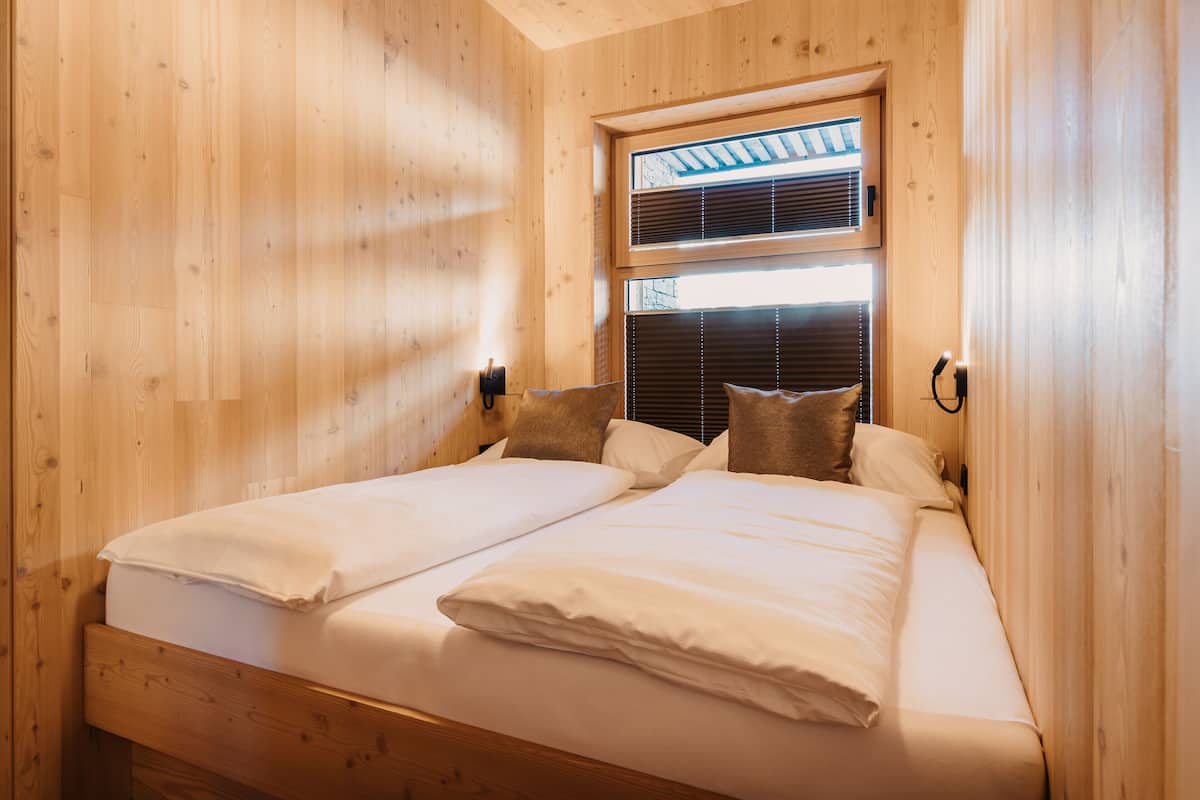 A double bed with white linens in a wooden cabin room with a window and blinds.