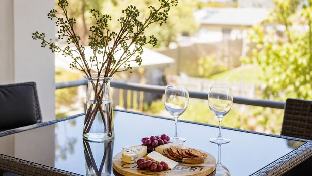 A glass-top table with a cheese board, grapes, and two wine glasses, set outdoors with a wicker chair and a vase with branches. - Seaview Cowes by Ready Set Stay (Cowes)