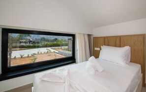 Quarto de hotel com uma janela grande, uma cama bem arrumada e vista para um edifício e vegetação lá fora.