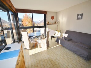 A living room with a sofa, a dining table with chairs, a kitchen area, and large windows offering a view of trees and mountains.