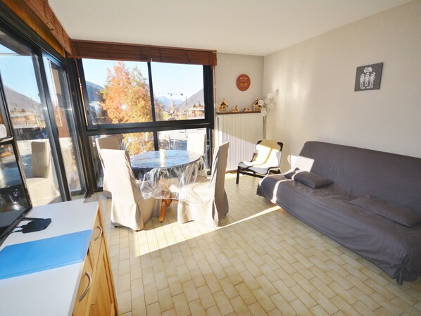 A living room with a sofa, a dining table with chairs, a kitchen area, and large windows offering a view of trees and mountains.
