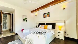 Une chambre à coucher moderne avec un grand lit, des tables de chevet, une petite table, un ventilateur de plafond et une porte avec un panneau de verre.