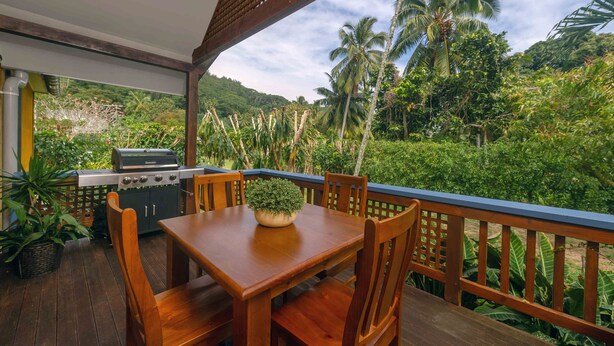 A wooden deck with a table and chairs, a grill, and a view of a lush green landscape with palm trees.