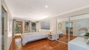 A modern bedroom with a large bed, a bedside table with a lamp, a glass door leading to a bathroom, and a view of the outdoors. - Pretty Beach Bay Vista by HolidayCo. (PRETTY BEACH)