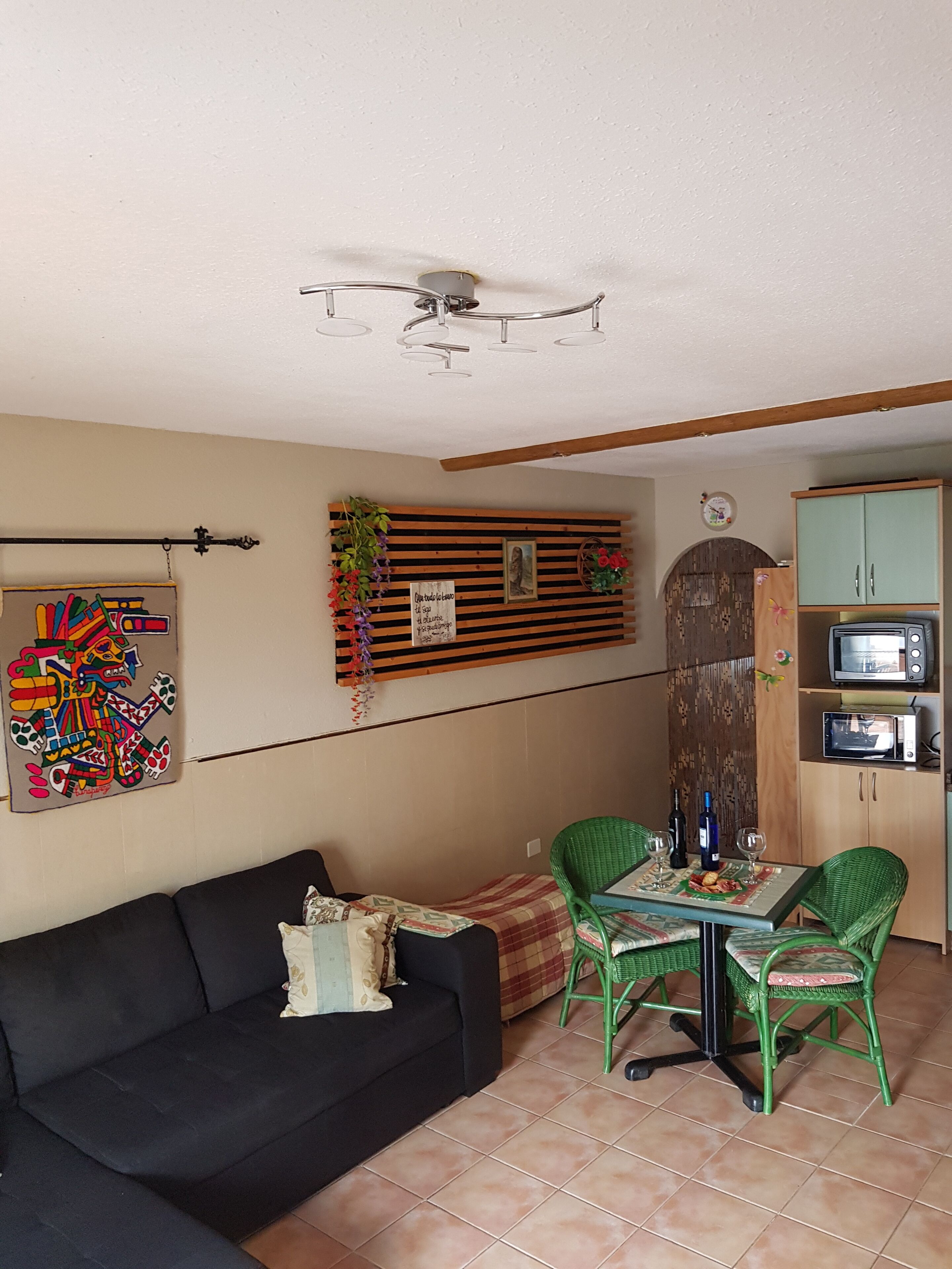 A cozy living area with a black sofa, green wicker chairs, a small dining table, and a kitchen area with a microwave and cabinets.