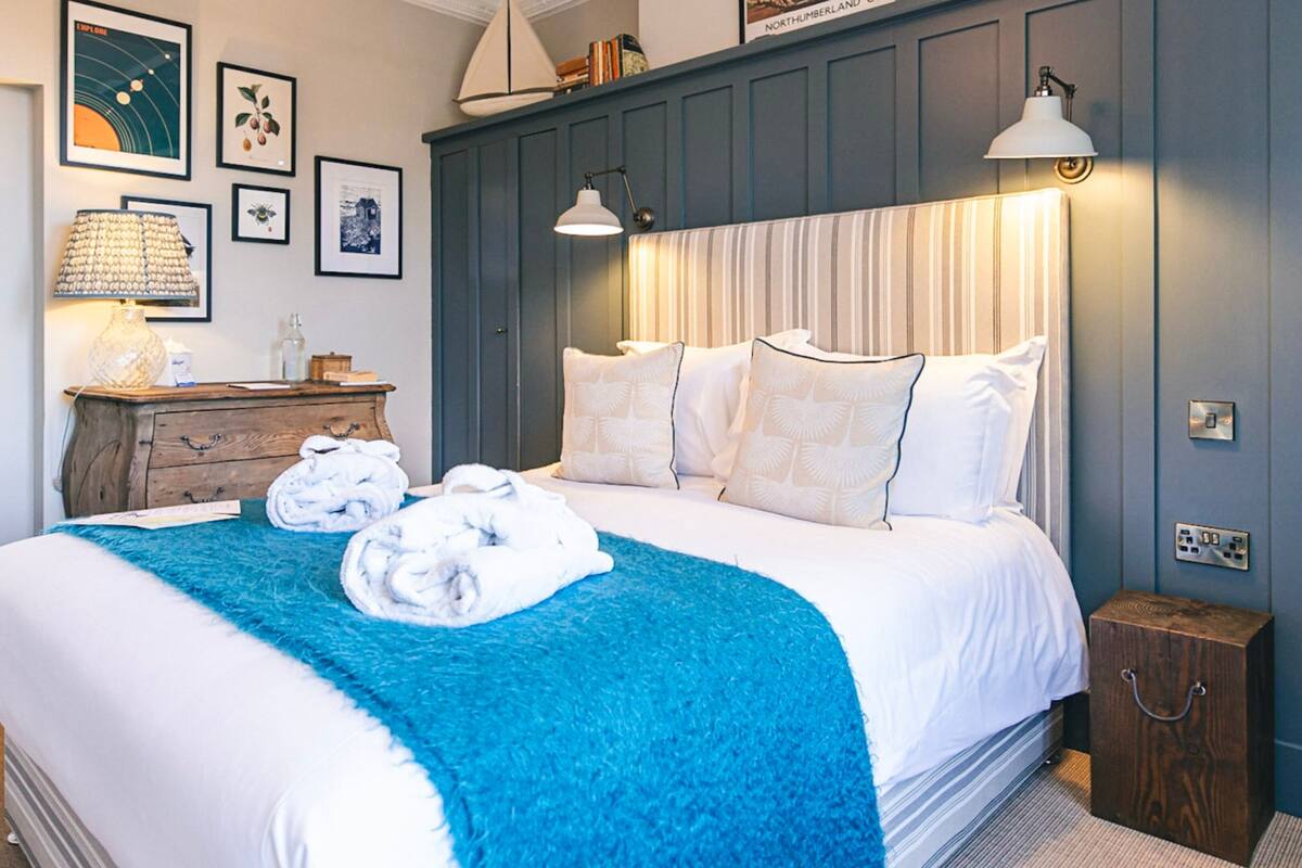 A neatly made bed with white linens and a blue throw blanket, a wooden dresser with a lamp, and framed pictures on the wall.