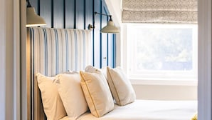 A bedroom with a striped headboard, two bedside lamps, and a window with patterned curtains. - The Whittling House (Alnwick)