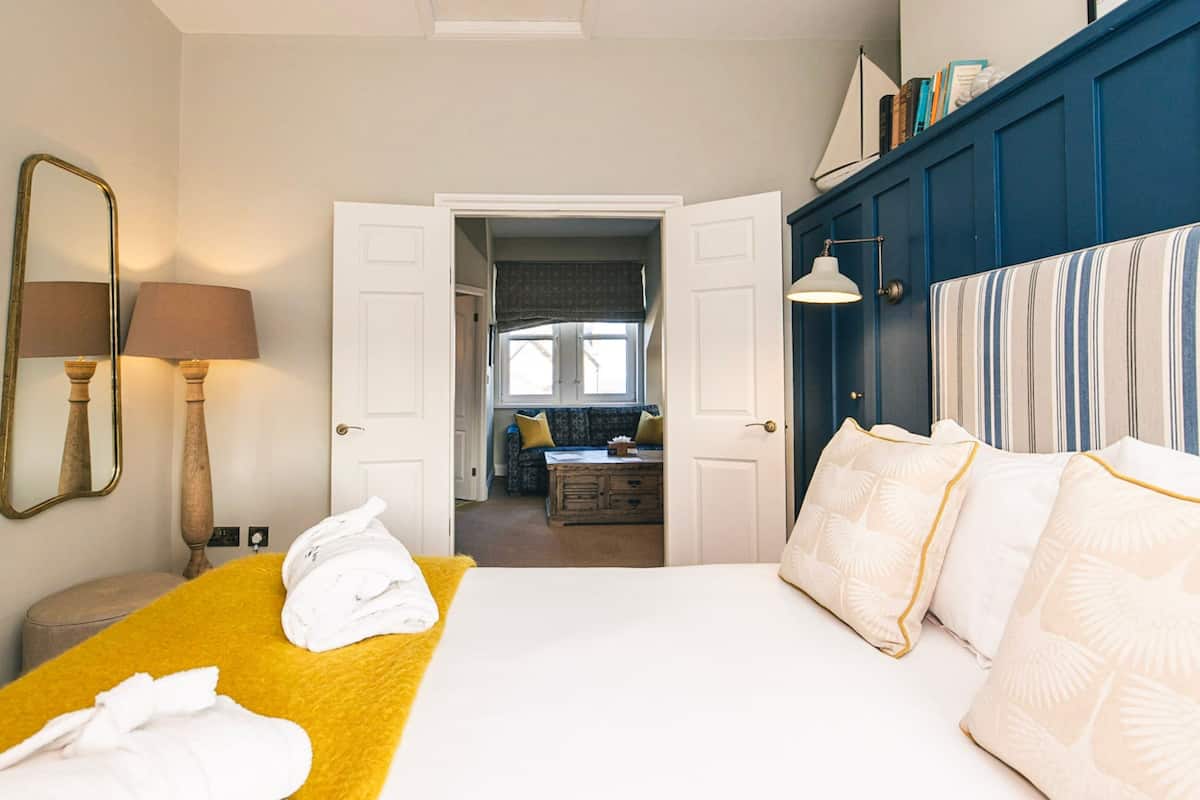 A hotel room with a bed, a yellow throw, a mirror, and a view into the adjacent room.
