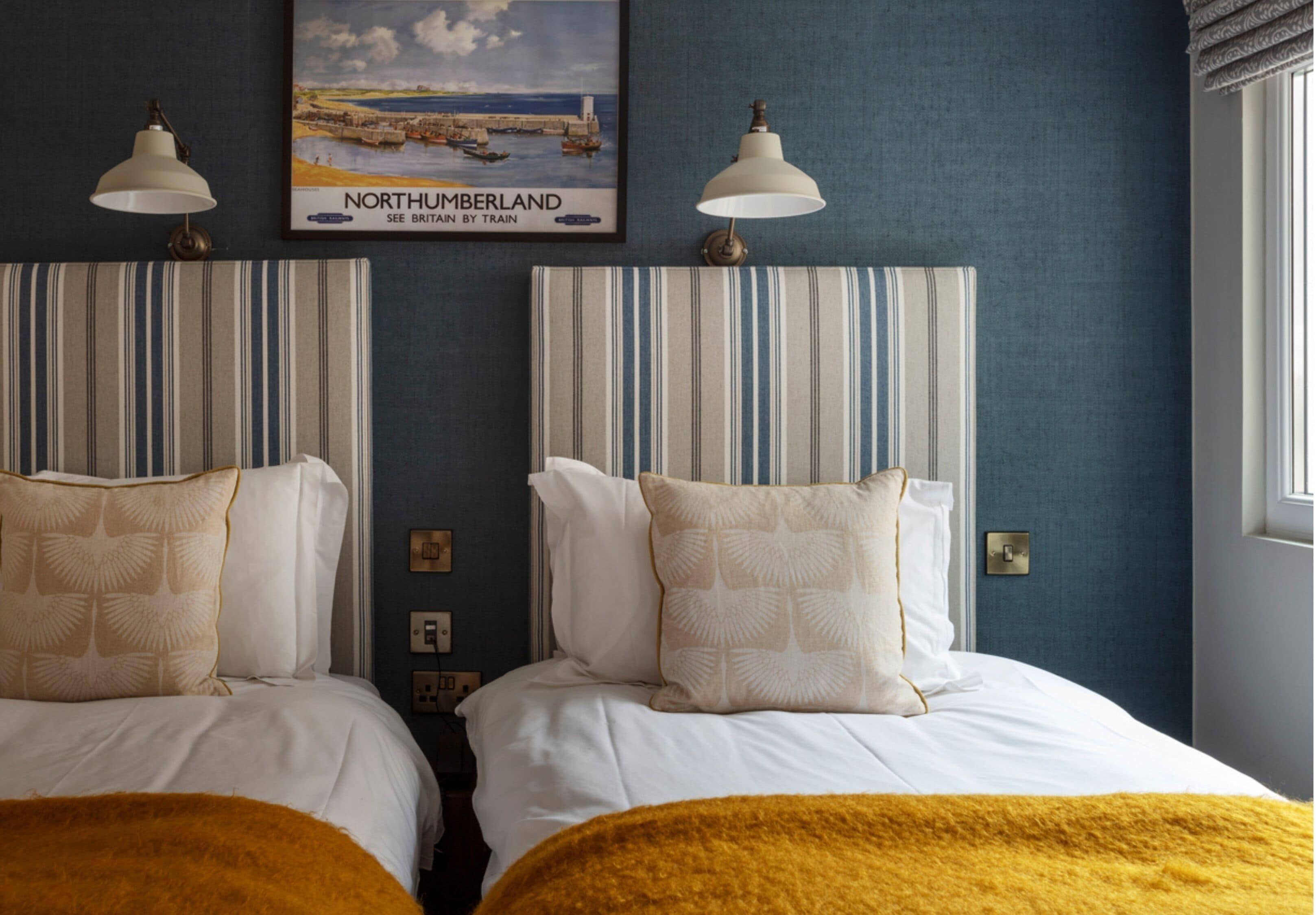 A hotel room with two beds, a framed picture of Northumberland, and a window on the right.