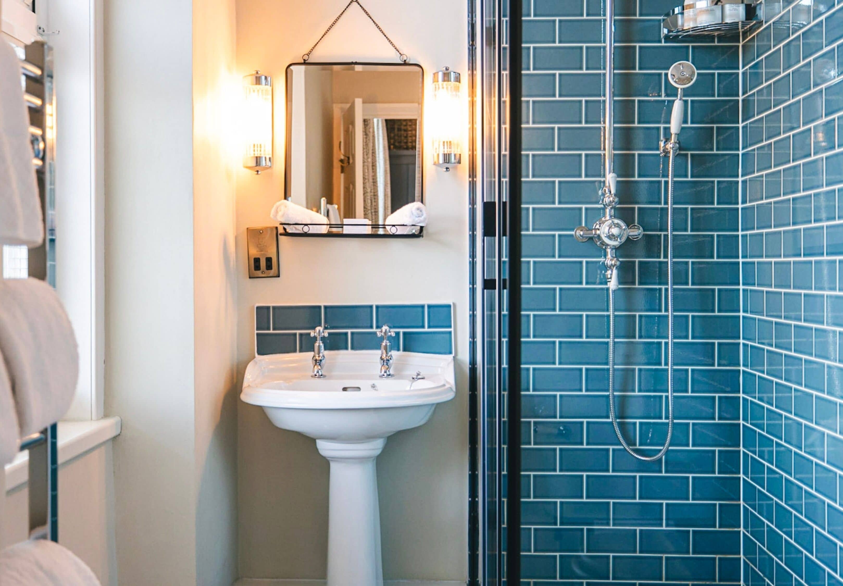 A bathroom with a white pedestal sink, a mirror with wall sconces, and a shower with blue-tiled walls.