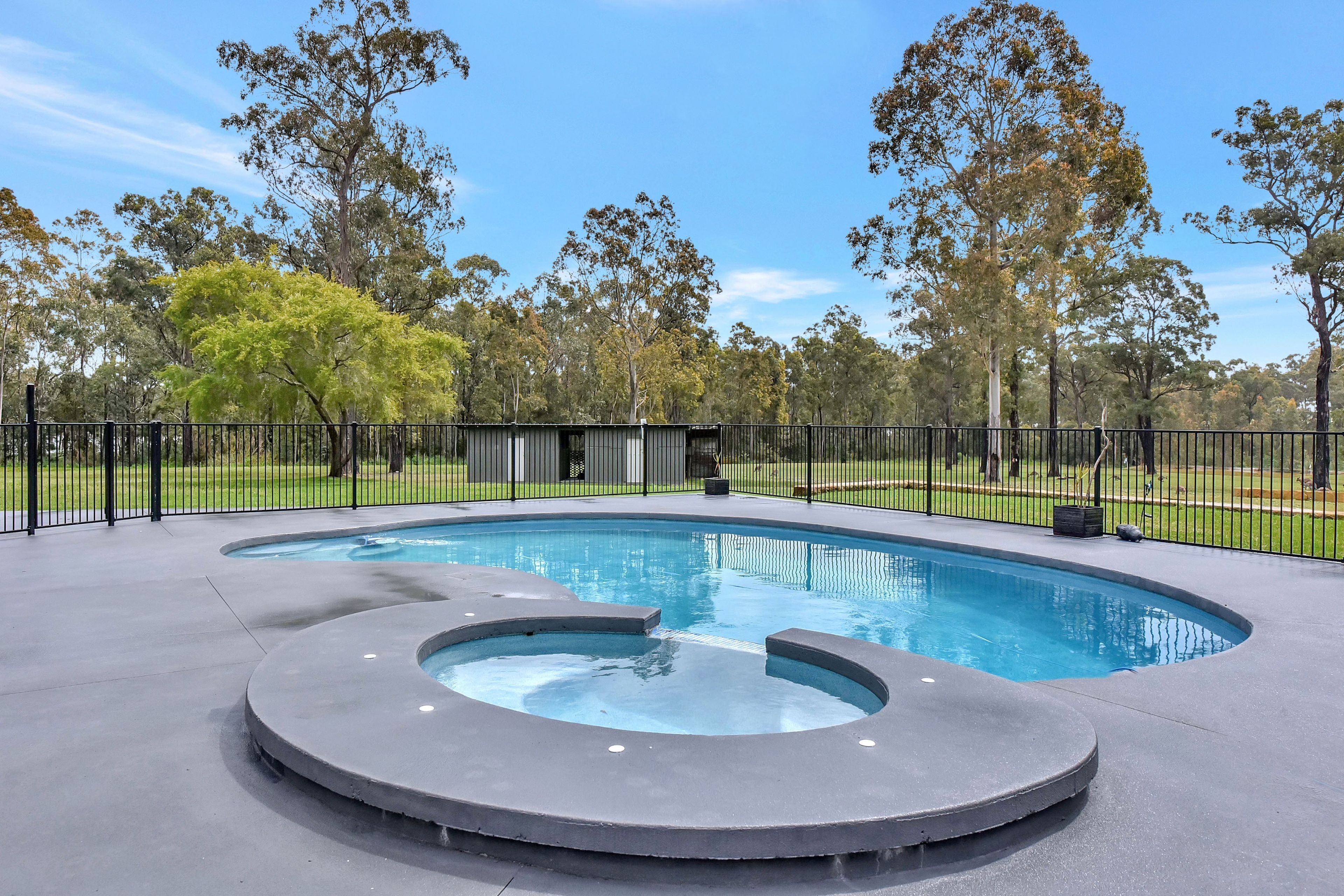 Une piscine avec un jacuzzi circulaire au centre, entourée d’une clôture et d’arbres.